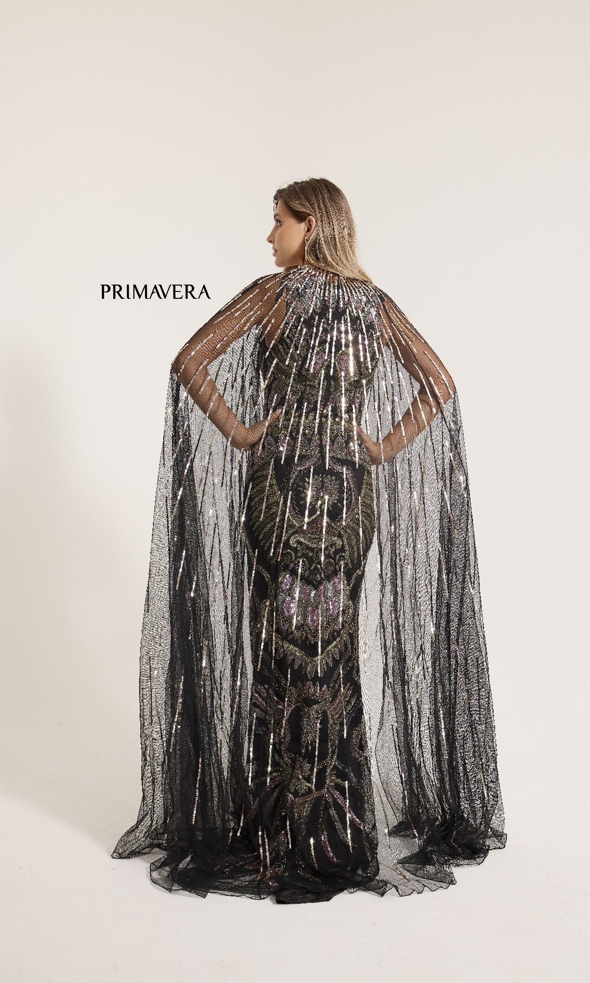 A woman faces away in a sleeveless, black, floor-length Primavera 12177 formal dress adorned with sequins and a sheer sparkling cape, standing before a plain white backdrop.